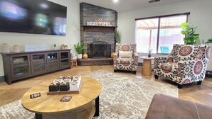 A living room with a fireplace, two patterned armchairs, a coffee table, and a TV mounted on the wall. - Zion Happy Retreat- Game Room, Sauna, & workspace! Pet Friendly! (Toquerville)