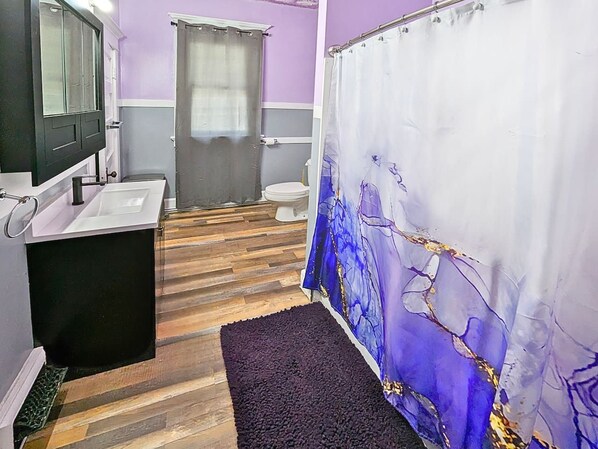 A bathroom with a wooden floor, a black bathtub, a white sink, and a shower curtain with a blue abstract design. - 6BR Crew House | Gym, 2 Washers, 55" TVs (Evansville)