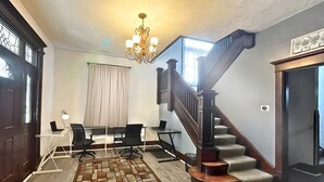 Ein geräumiger Raum mit einer Holztreppe, einer Chandelüster, einem Schreibtisch mit zwei Stühlen und einem großen Fenster.