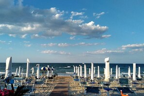 A beach with rows of sun loungers and umbrellas, a wooden walkway, and the ocean in the background.