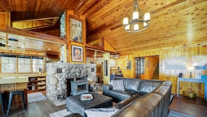 A cozy cabin interior with a stone fireplace, leather sofa, wooden walls, and a dining area.