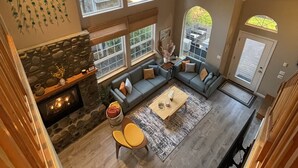 A spacious living room with a stone fireplace, large windows, and a high ceiling.