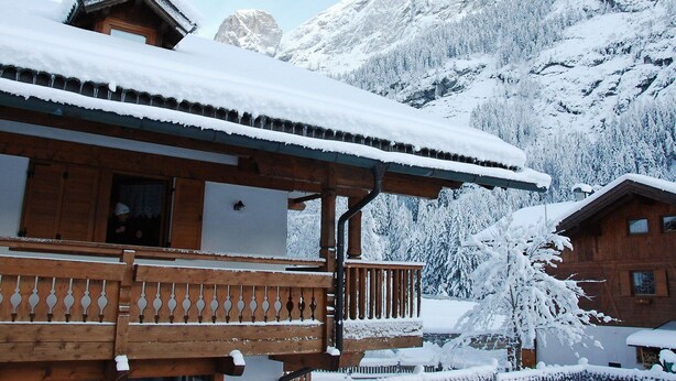Un chalet de madera con techo nevado y un balcón, rodeado de árboles cubiertos de nieve y montañas al fondo.