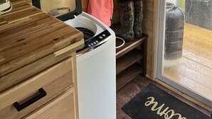 A compact room with a washing machine, a wooden cabinet, a television mounted on the wall, and a door leading outside. - Lavender Haze Cottage in the Woods (Oakland)