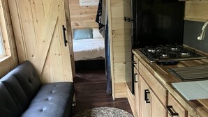 A compact, modern interior with a sofa, a round rug, wooden cabinets, a stove, and a bed in the background. - Lavender Haze Cottage in the Woods (Oakland)