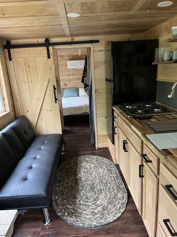 Un interior moderno y compacto con un sofá, una alfombra redonda, armarios de madera, una estufa y una cama al fondo.