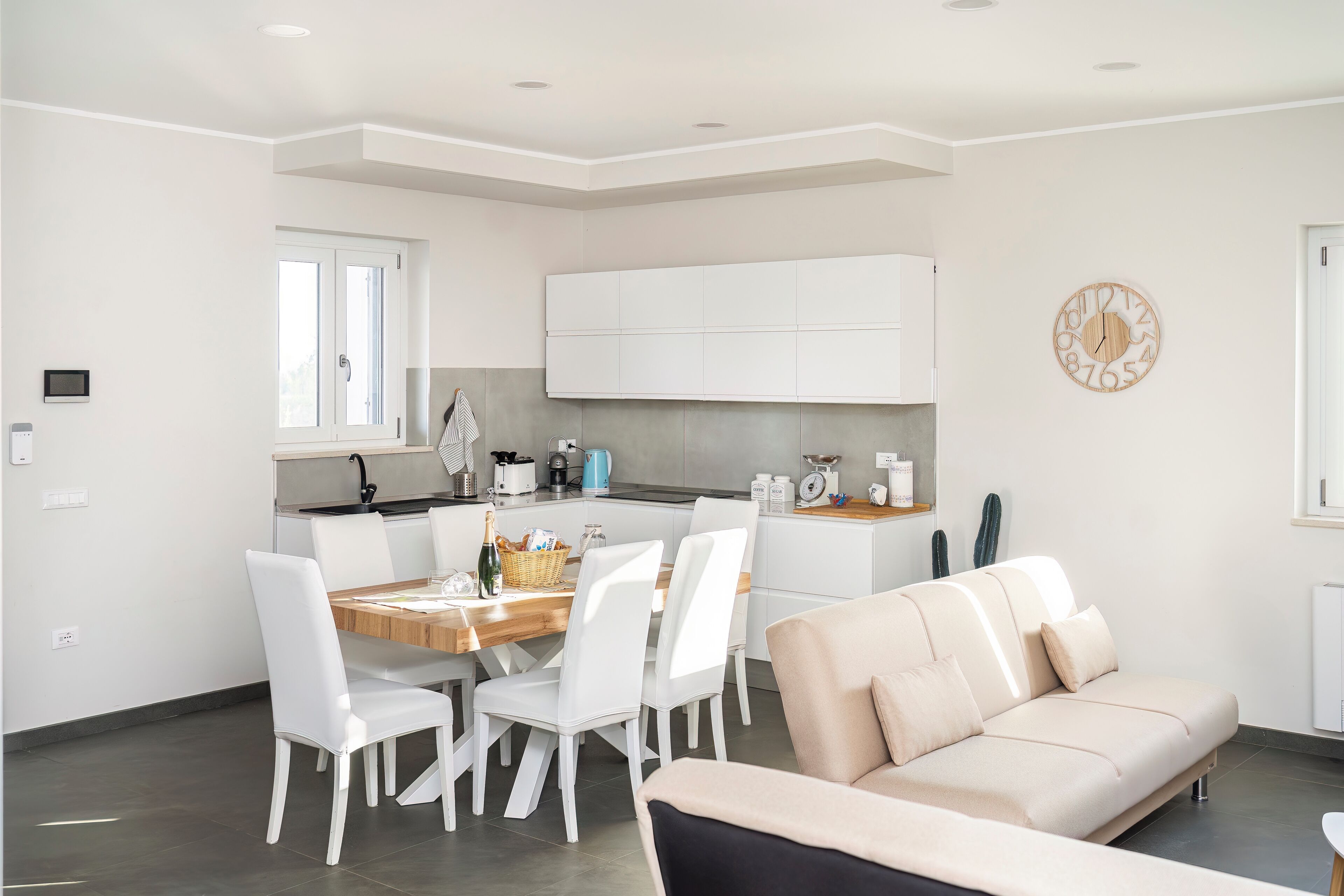 Una cucina moderna con mobili bianchi, un tavolo da pranzo con sedie e un divano beige in un soggiorno luminoso.