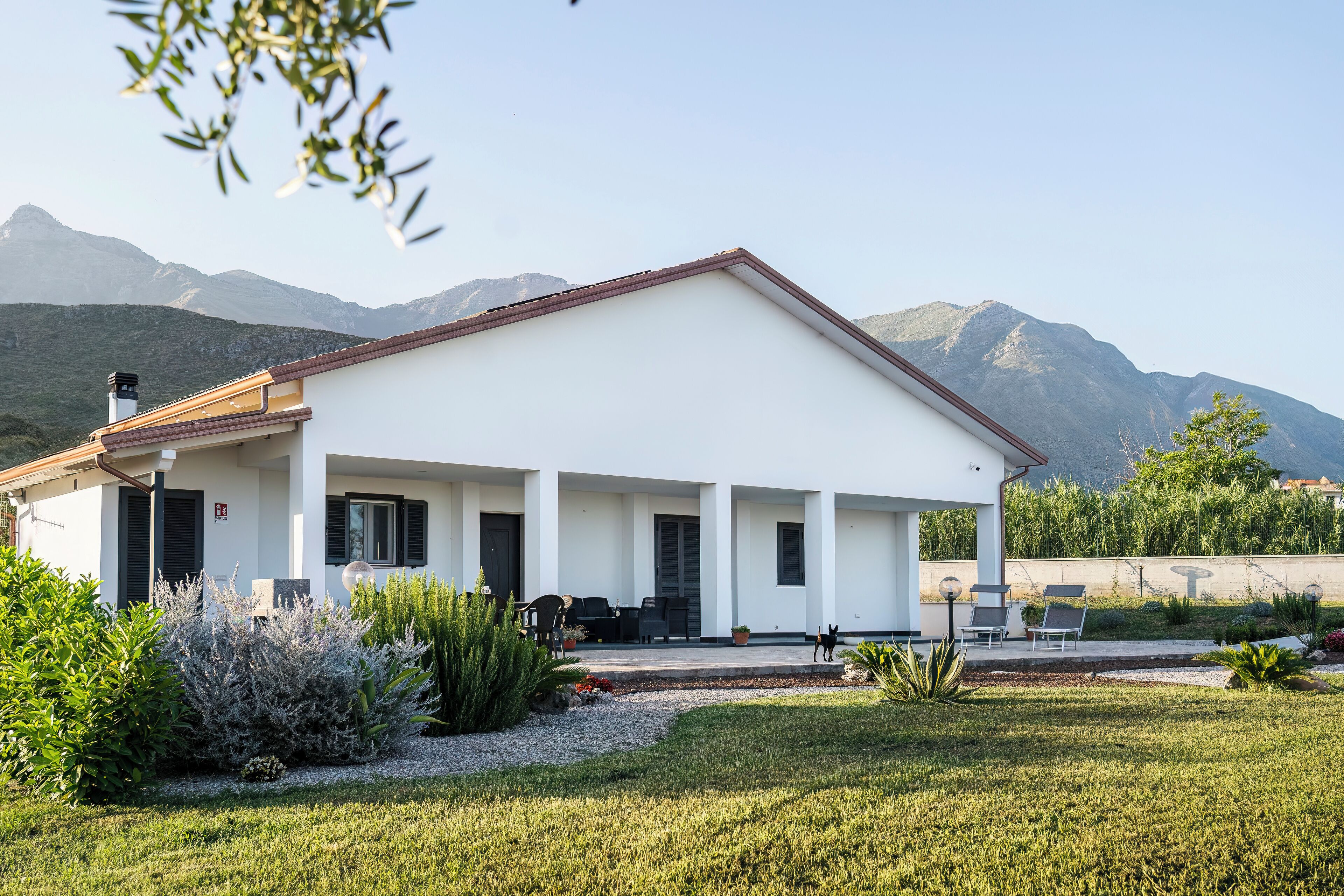 Un edificio moderno con tetto piatto e pareti bianche, circondato da un giardino curato e montagne sullo sfondo.