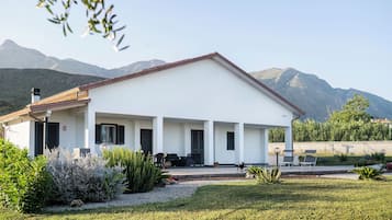Un edificio moderno con tetto piatto e pareti bianche, circondato da un giardino curato e montagne sullo sfondo.