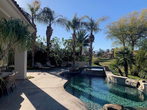 A swimming pool with clear water, surrounded by palm trees and a patio area with lounge chairs.