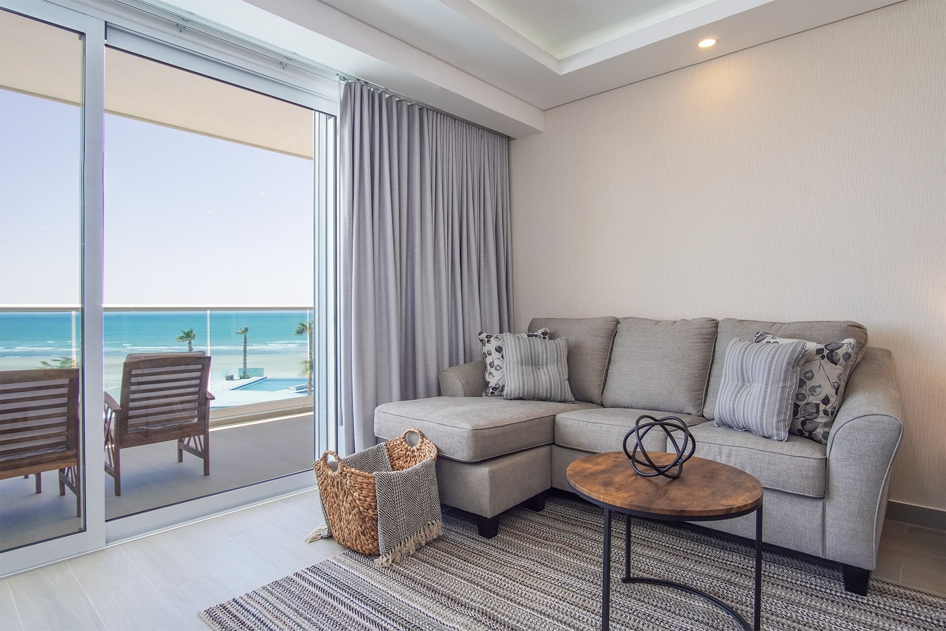 A modern living room with a grey sectional sofa, a small wooden coffee table, and a view of the beach through large sliding glass doors.