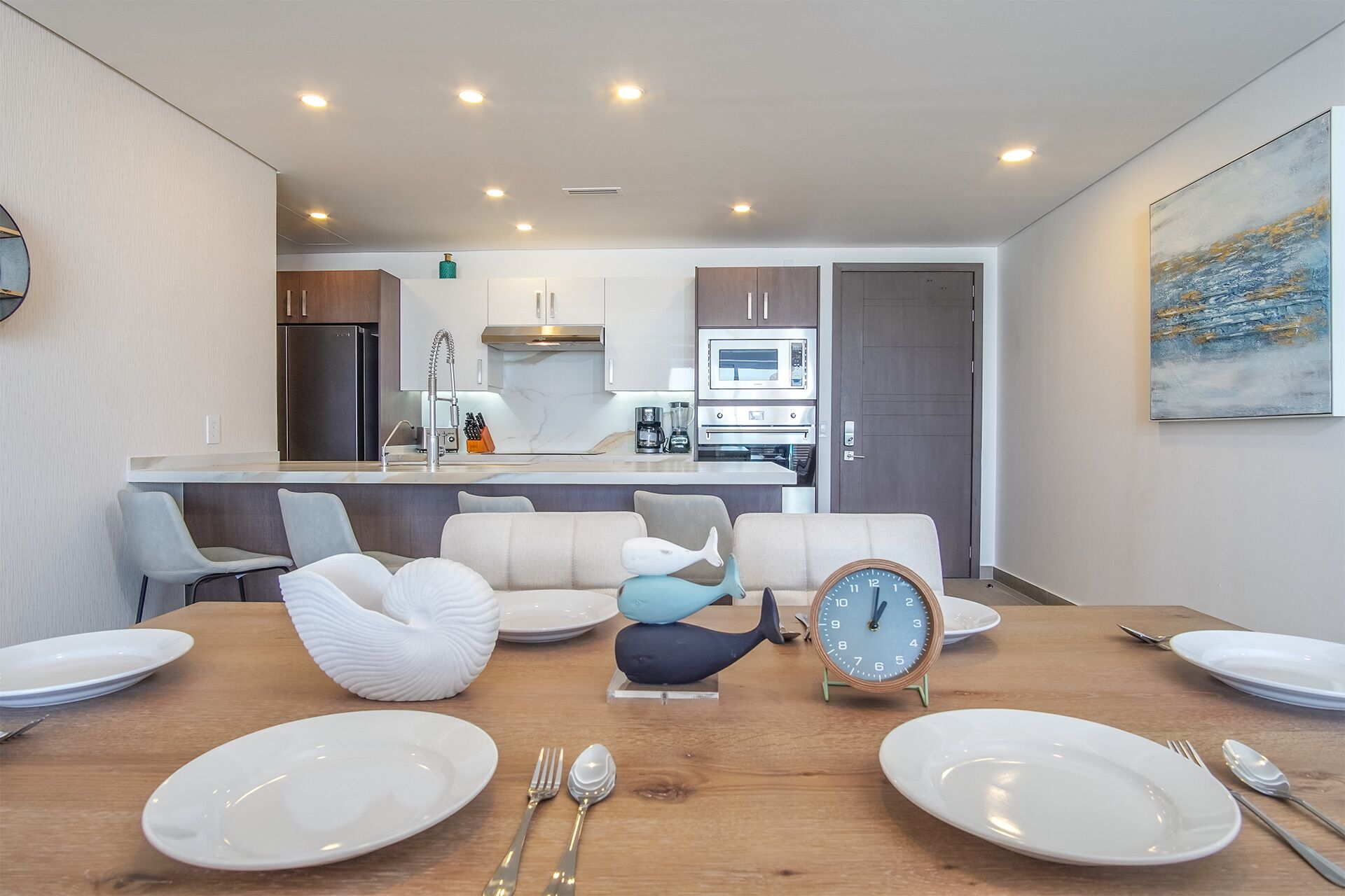 A modern kitchen with a central island, a dining area with a wooden table set for four, and a living area with a sofa and decorative items.