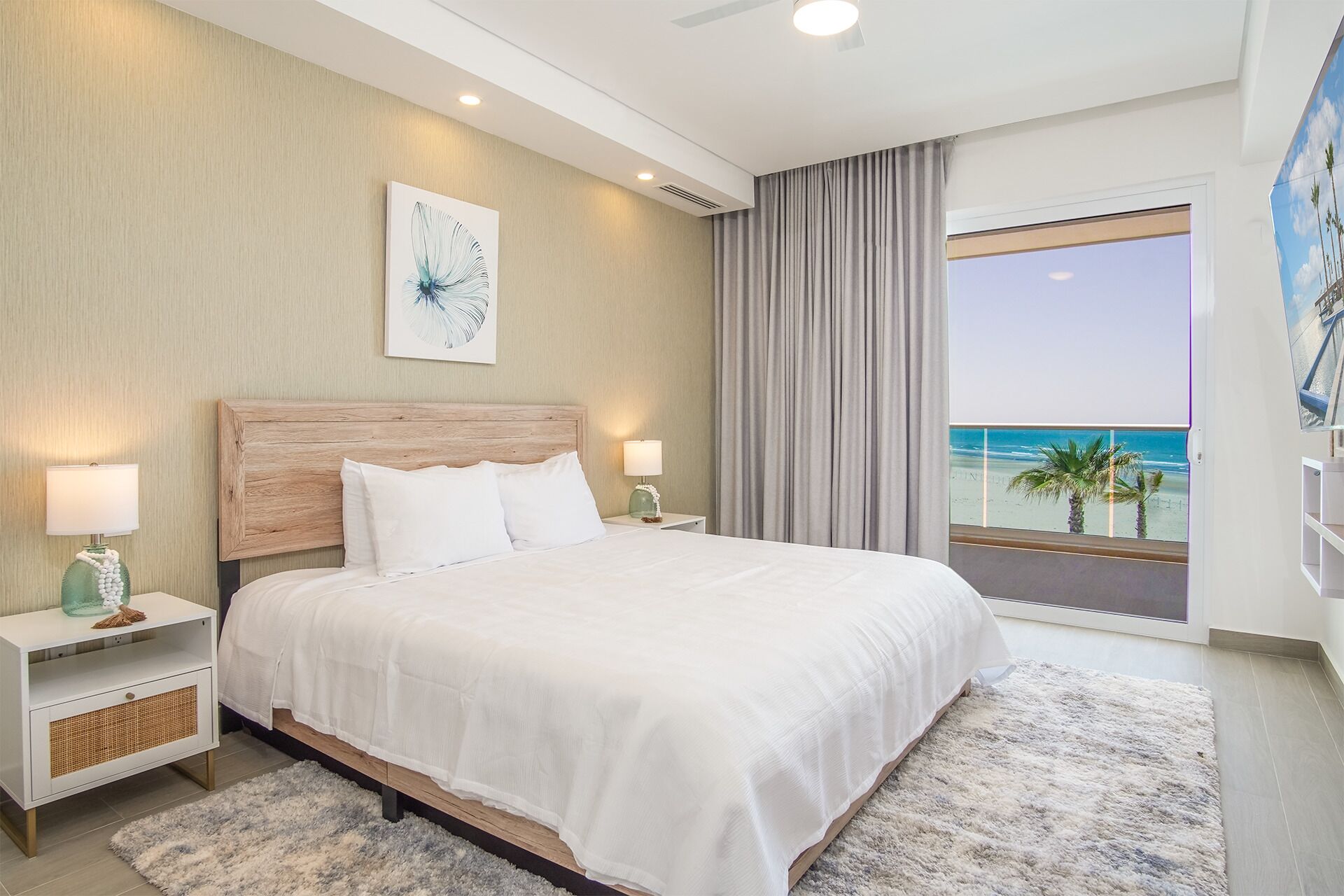 A modern hotel room with a large bed, two bedside tables with lamps, a view of the beach, and a ceiling fan.