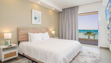 A modern hotel room with a large bed, two bedside tables with lamps, a view of the beach, and a ceiling fan.