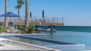 Zona de piscina con sillones de descanso, sombrillas y palmeras.