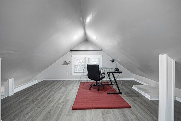 An attic room with a sloped ceiling, a black office chair, a desk with a lamp, and a red rug. - Spacious 4BD 2BA Home Near OSU (Columbus)