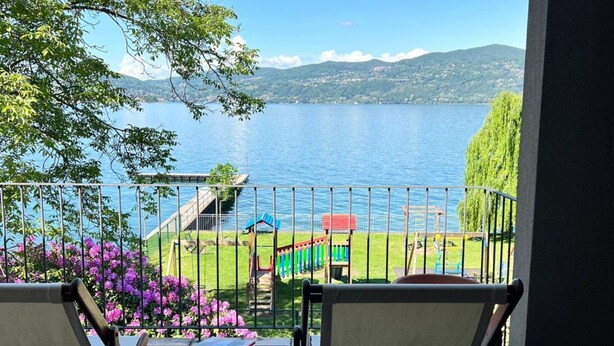 A balcony with two chairs, a table, and a view of a lake with mountains in the background. - Bnbook Casa Luccio Vista lago-Ispra (ISPRA)