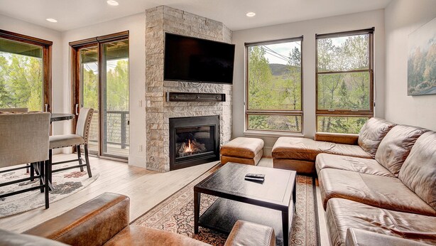 A modern living room with a stone fireplace, leather sofas, a flat-screen TV, and large windows offering a view of trees. - Blue River Flats in Silverthorne, amazing location and panoramic mountain views (Silverthorne)