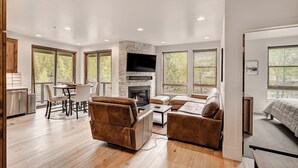 A spacious living area with a stone fireplace, wooden flooring, and large windows offering views of trees. - Blue River Flats in Silverthorne, amazing location and panoramic mountain views (Silverthorne)