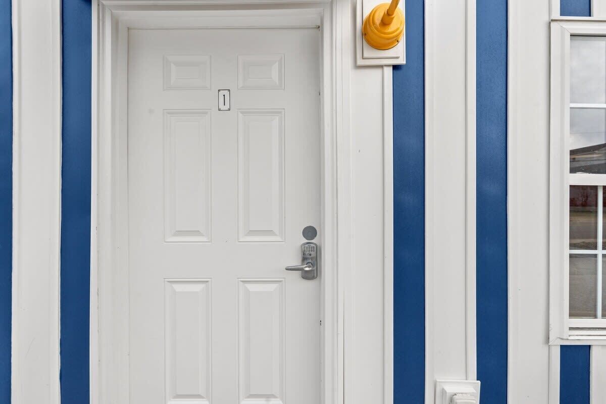 Une porte blanche dotée d’un clavier pour l’ouverture, un petit objet jaune accroché au mur et une fenêtre à cadre blanc.