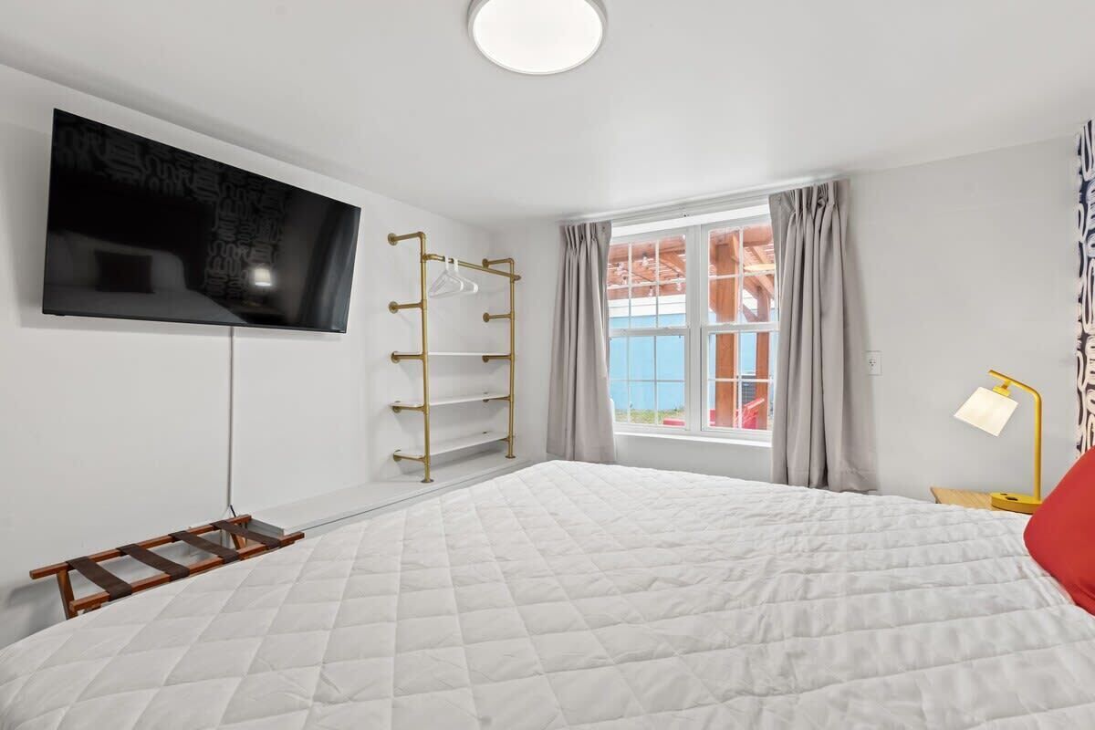Un dormitorio con una cama grande, un televisor montado en la pared, una ventana con cortinas y una lámpara de noche.