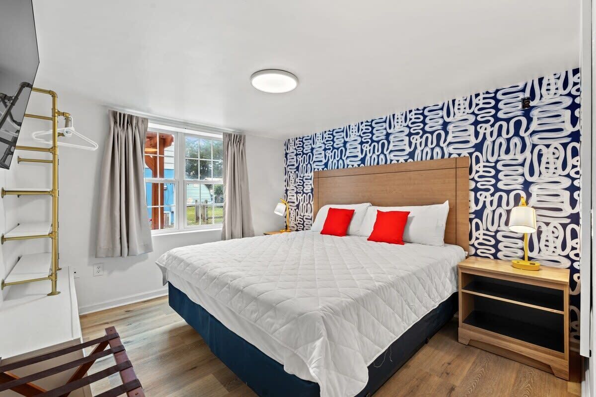 Una habitación de hotel con una cama grande, cabecera de madera, mesita de noche con lámpara, ventana con cortinas y papel tapiz con diseño en la pared.
