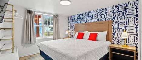 Una habitación de hotel con una cama grande, cabecera de madera, mesita de noche con lámpara, ventana con cortinas y papel tapiz con diseño en la pared.