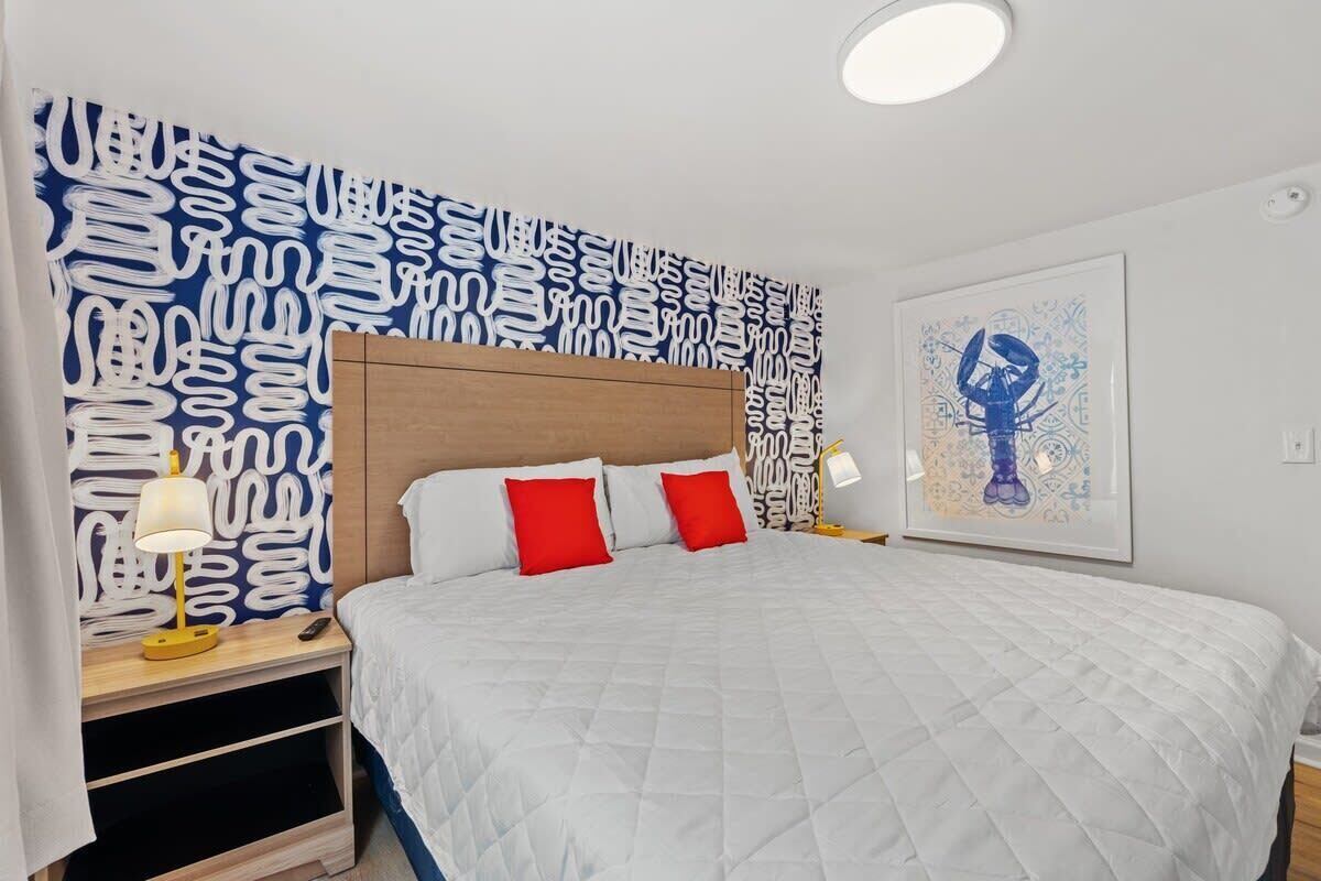 A bedroom with a large bed, two red pillows, a white quilted bedspread, a wooden headboard, a nightstand with a lamp, and a wall with a blue abstract pattern.