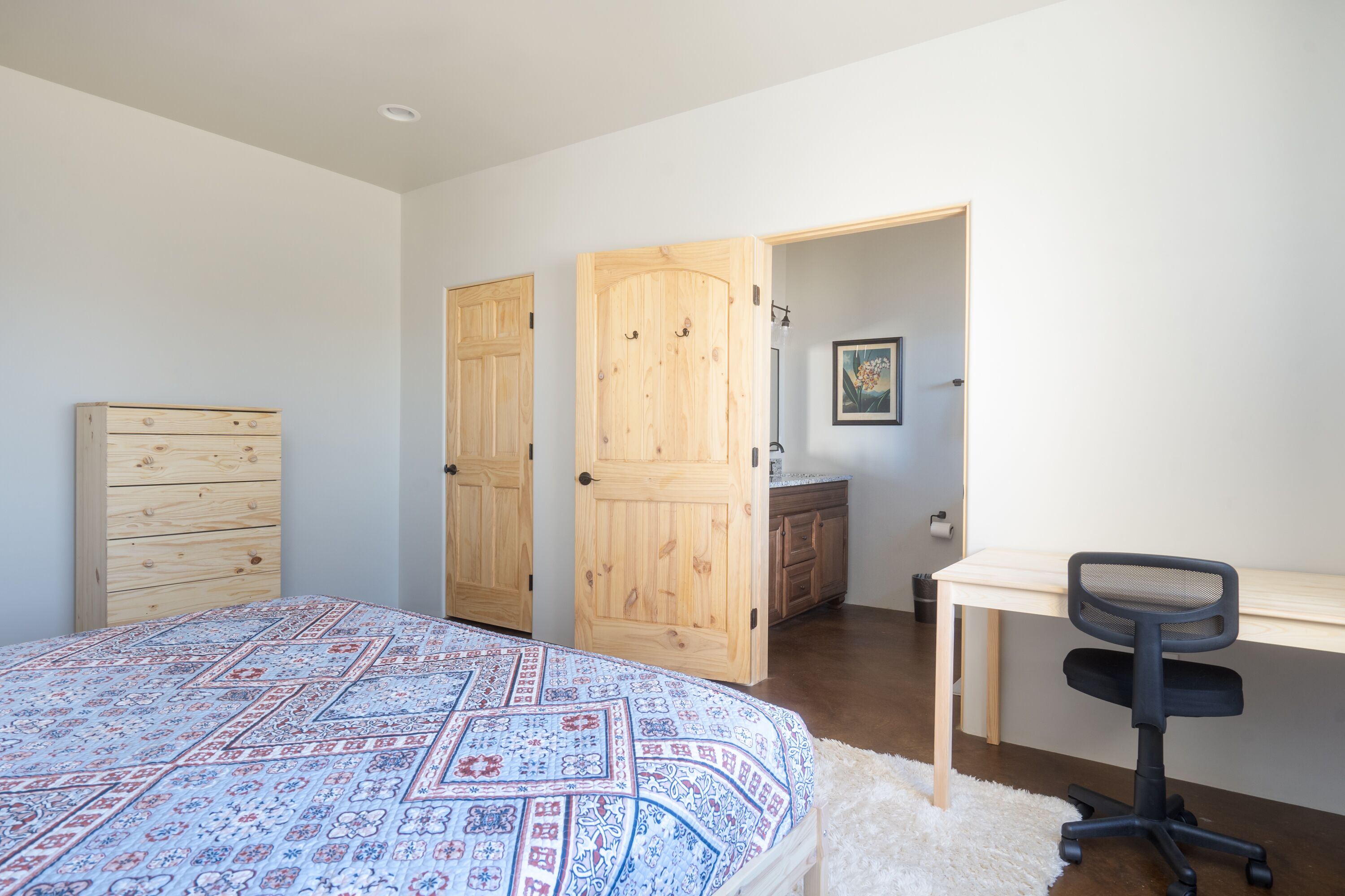 Un dormitorio con una cama, un aparador de madera, un escritorio con una silla y una puerta que da a otra habitación.