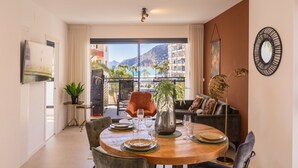 A dining area with a round table set for four, a sofa, and a view of the sea and mountains through a balcony.