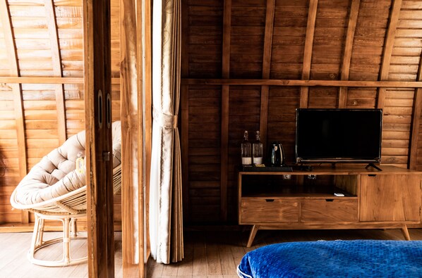 A wooden room with a television on a wooden cabinet, a wicker chair, and a bed with a blue blanket. - Kabinku Bali (Baturiti)