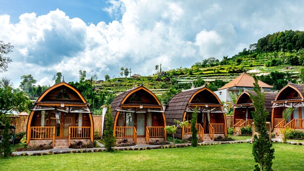A row of rustic, arched cottages with wooden balconies, set against a lush green landscape with terraced fields in the background.