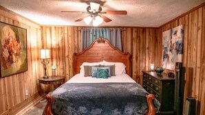 Un dormitorio con paneles de madera, una cama, una mesita de noche, un aparador, un ventilador de techo y un cuadro.