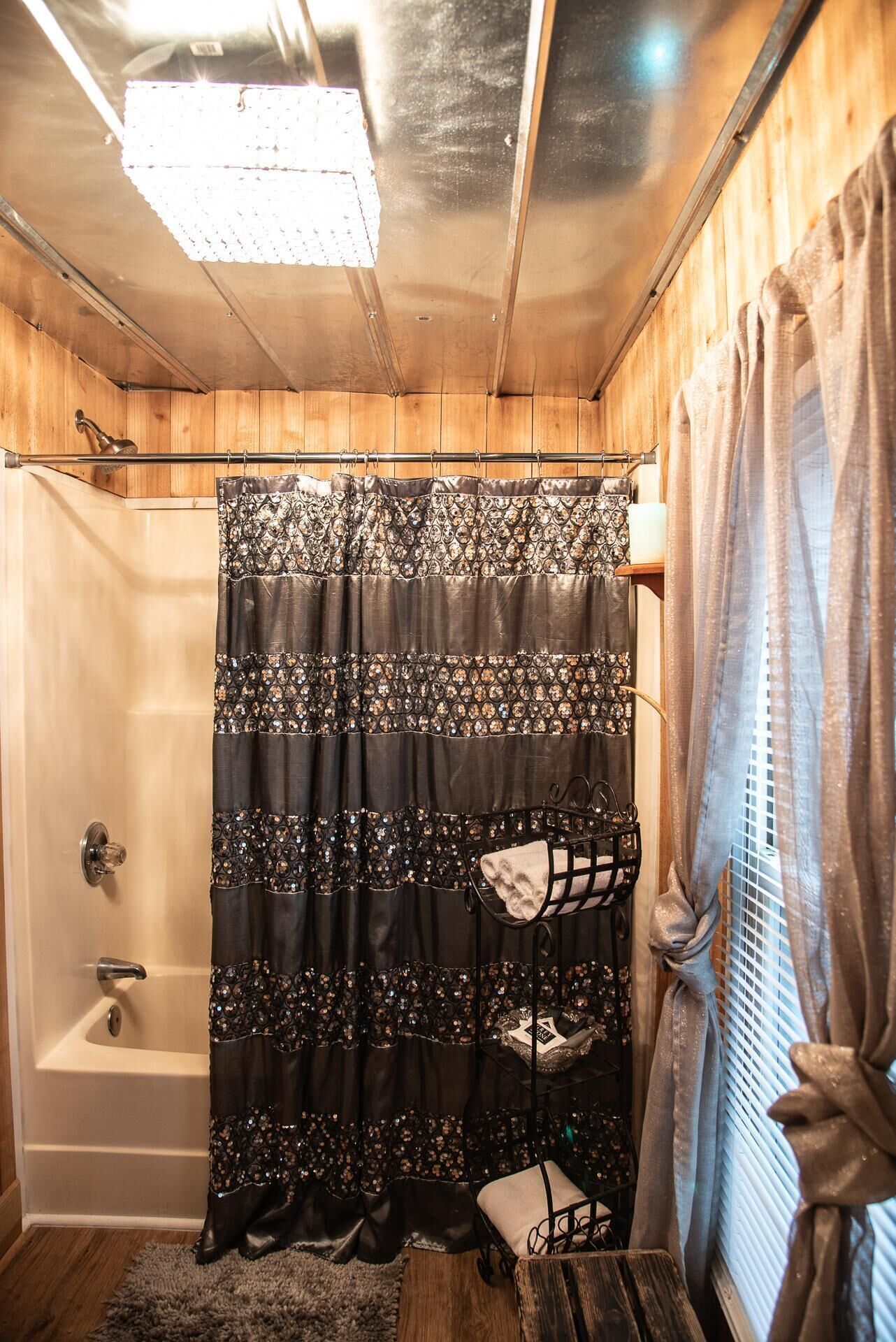 A bathroom with a shower curtain, a bathtub, a window with blinds, and a shelf with a towel.