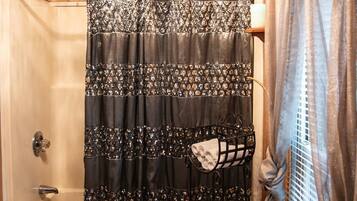 A bathroom with a shower curtain, a bathtub, a window with blinds, and a shelf with a towel.