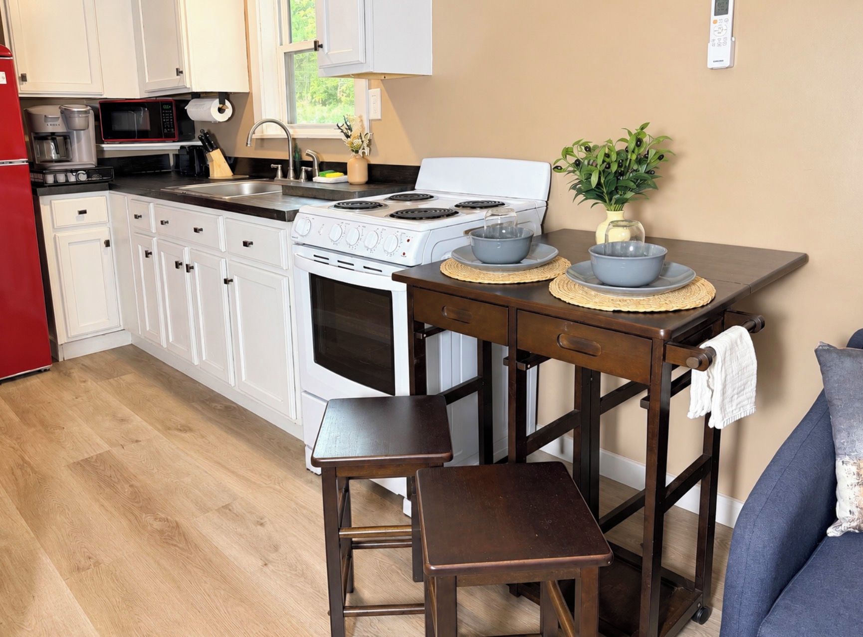 Une cuisine compacte avec des meubles blancs, une cuisinière, un four à micro-ondes et une machine à café. On y trouve une petite table à manger avec une chaise et une plante en pot.