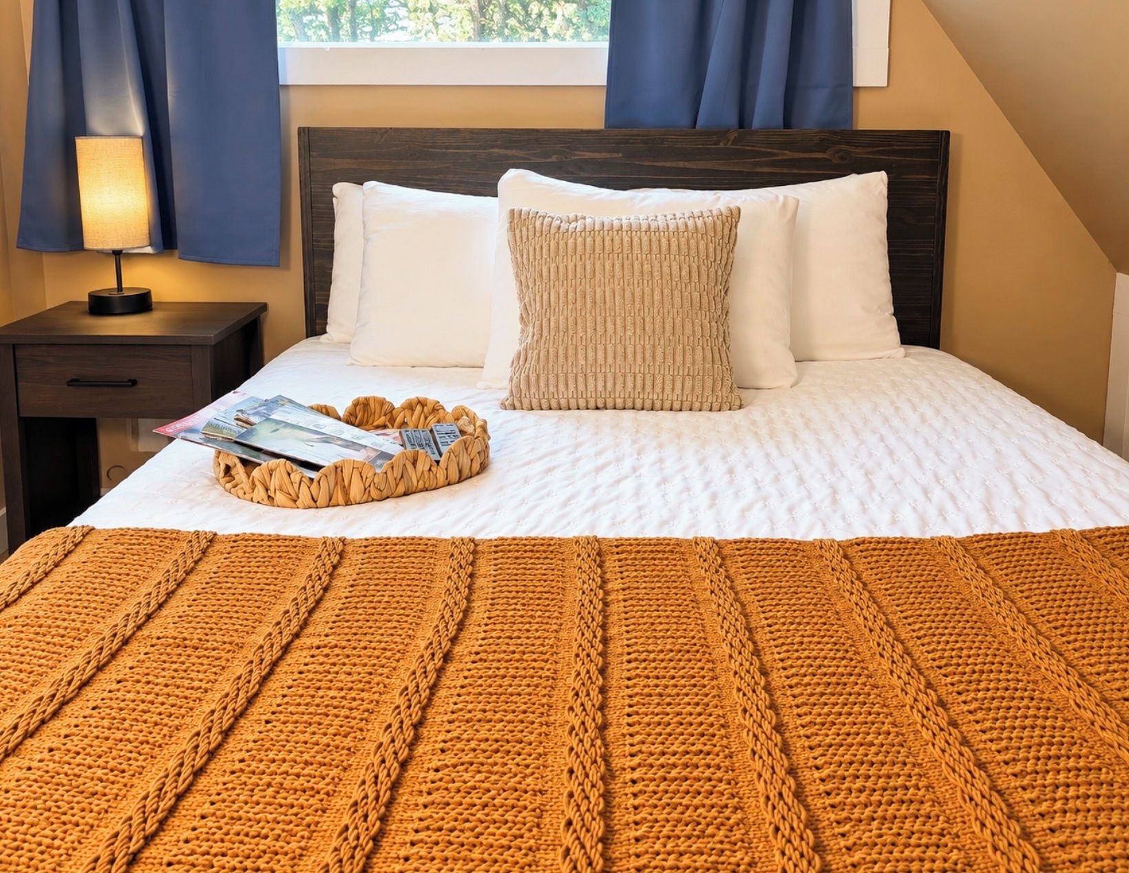 Un lit bien fait, avec une couette blanche, un plaid marron en tricot et un magazine posé sur un plateau en osier.