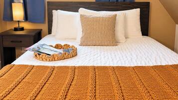 Un lit bien fait, avec une couette blanche, un plaid marron en tricot et un magazine posé sur un plateau en osier.