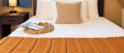 Un lit bien fait, avec une couette blanche, un plaid marron en tricot et un magazine posé sur un plateau en osier.