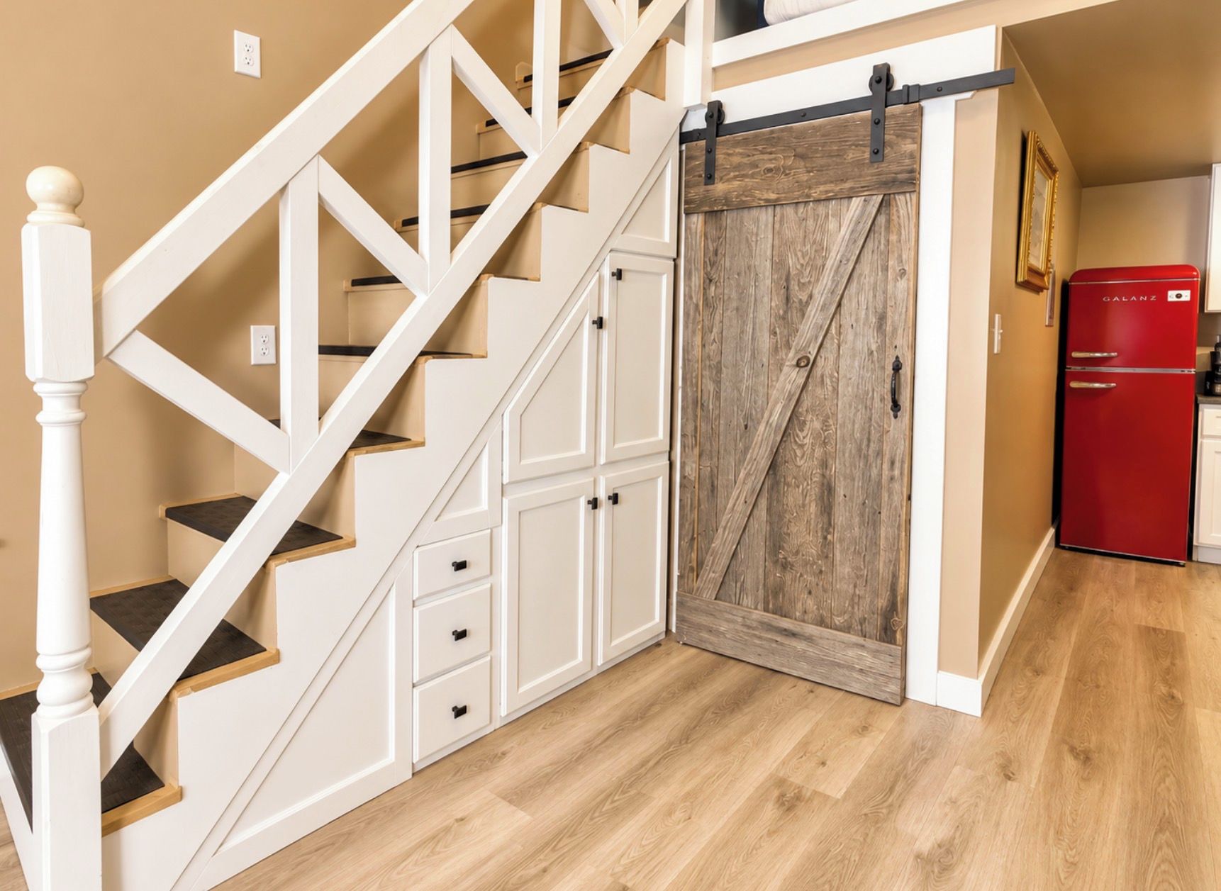 Un escalier avec des rampes blanches et des marches en bois, menant à une pièce dotée d’une porte en bois et d’un appareil électroménager rouge.