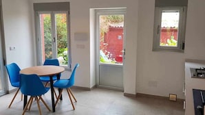 A dining area with a round table and blue chairs, two windows, and a red wall visible through the windows. - Bungalow n°6 on the Lighthouse Road (Lariño)