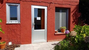 A red building with a white door and windows, potted plants on the windowsill, and a small garden area. - Bungalow n°6 on the Lighthouse Road (Lariño)