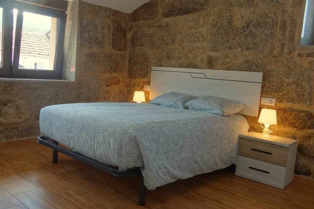 Un dormitorio con pared de piedra, una cama con cubrecama estampado, dos mesitas de noche con lámparas y una ventana con persianas.