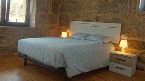 Une chambre avec un mur en pierre, un lit recouvert d’une couvre-lit à motifs, deux tables de chevet avec des lampes et une fenêtre avec des stores.