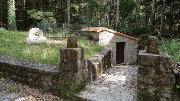 Un chemin de pierres mène à un petit bâtiment en pierre surmonté d’un toit en tuiles, l’ensemble étant entouré d’un mur de pierres et d’une clôture.