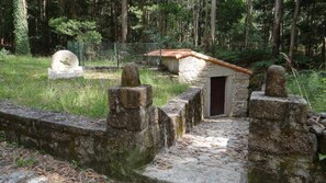 Un chemin de pierres mène à un petit bâtiment en pierre surmonté d’un toit en tuiles, l’ensemble étant entouré d’un mur de pierres et d’une clôture.