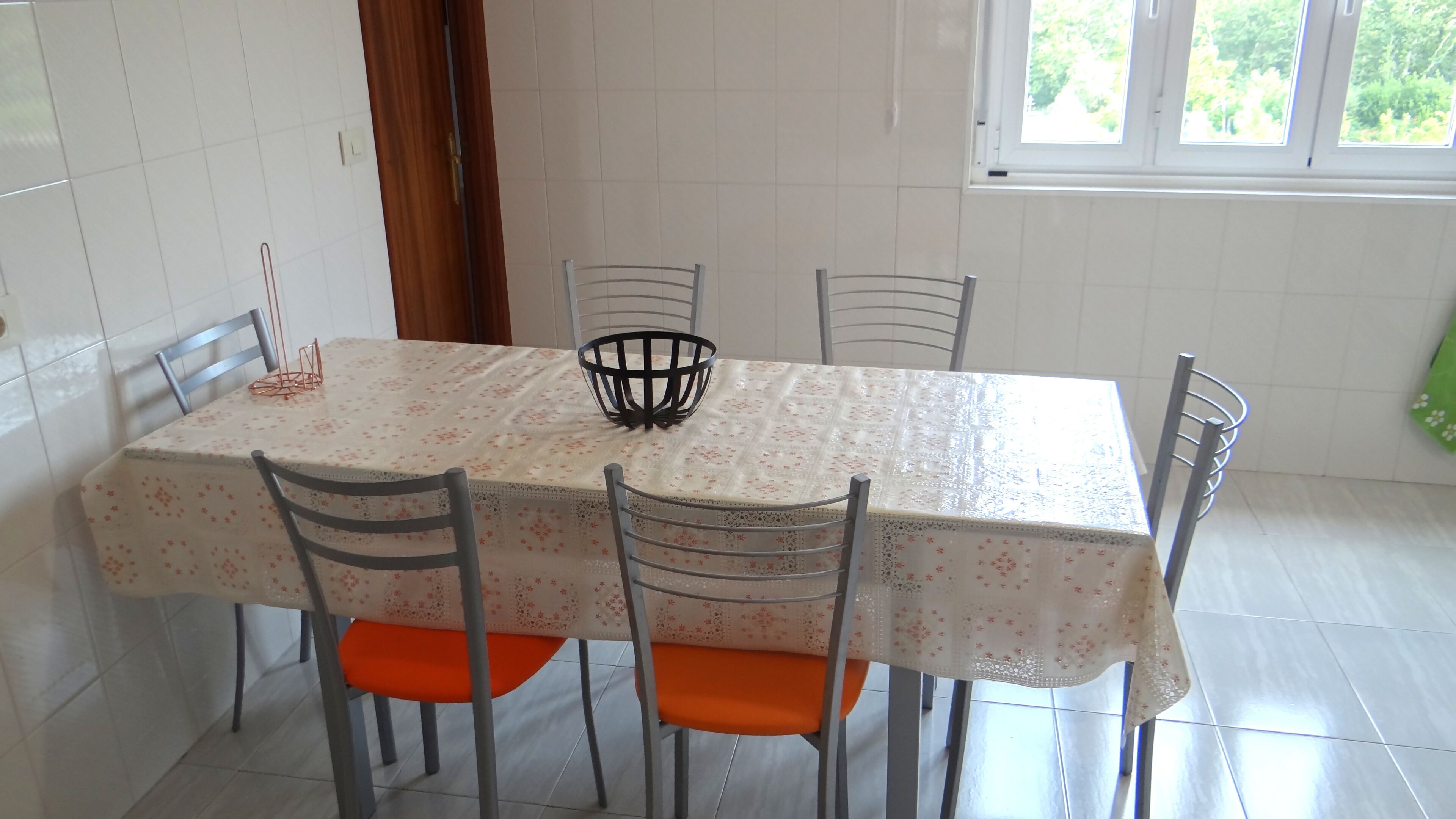 Un coin repas avec une table recouverte d’une nappe à motifs floraux, quatre chaises dotées de coussins orange et un panier noir posé sur la table.
