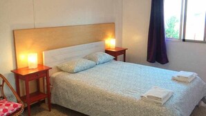 Un llit ben fet amb un edredó estampat, dues làmpades de nit, una tauleta de nit de fusta i una finestra amb cortina.