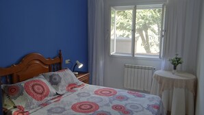 Un dormitorio con cama, mesita de noche, lámpara, ventana con cortinas, radiador y una pequeña mesa con un florero.