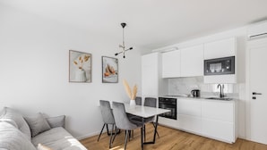 A modern kitchen with white cabinets, a microwave, and a sink. There is a dining area with a white table and grey chairs. - Did' (Split)